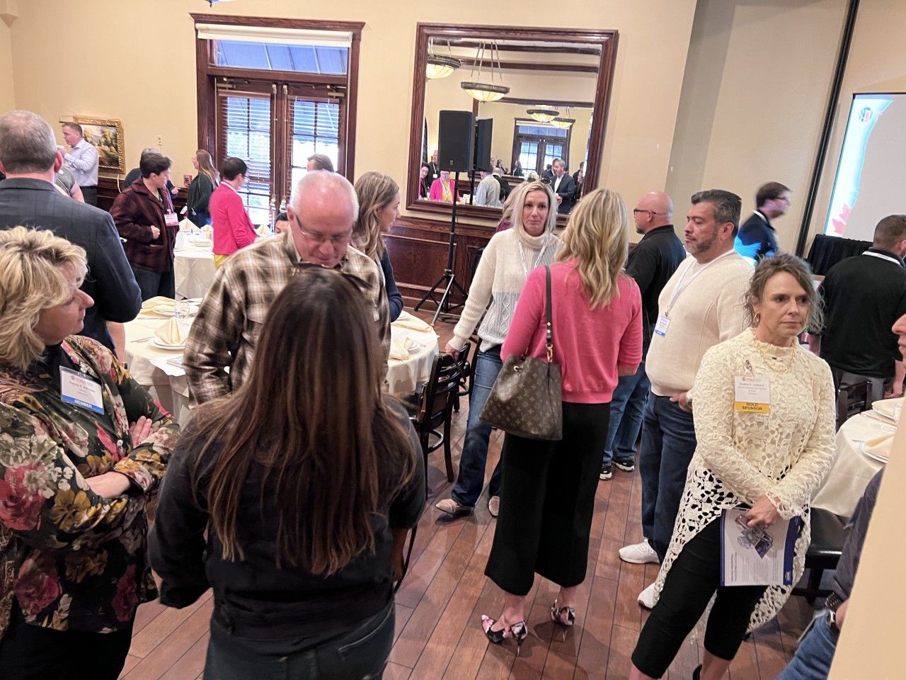 People mingling in a room with round tables, mirrors, and a screen. Some hold drinks, and a few wear name tags.