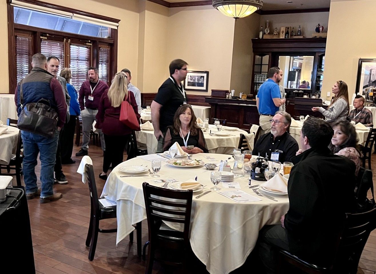 People at tables and standing in a restaurant, socializing. Some are eating, others talking. Neutral lighting.