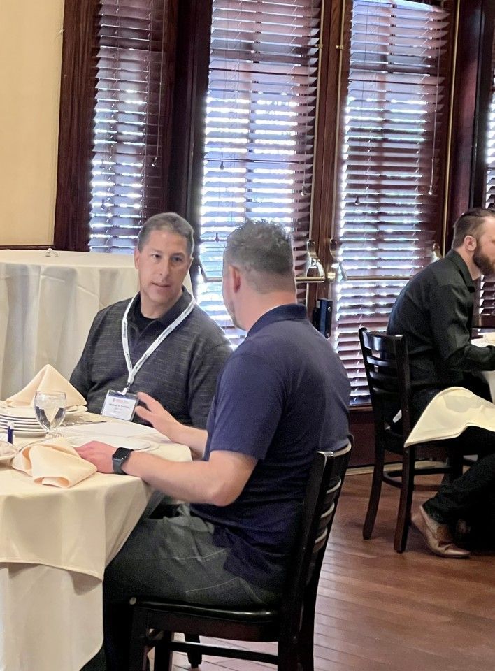 Two men at a table, conversing in a restaurant setting. One looks at the other, who's turned partially.