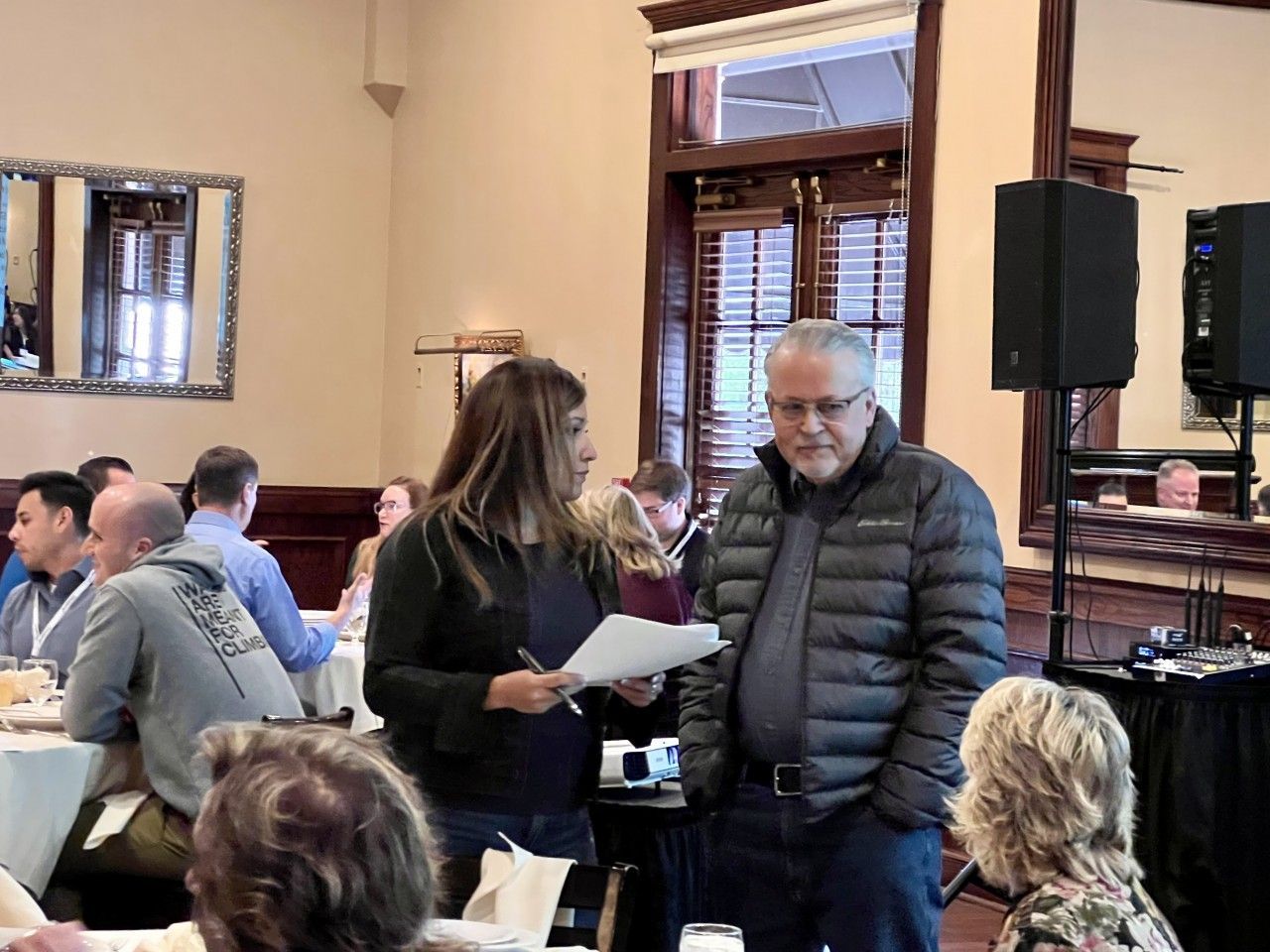 Woman with papers talking to a man in a quilted jacket, in a restaurant with tables and other people.