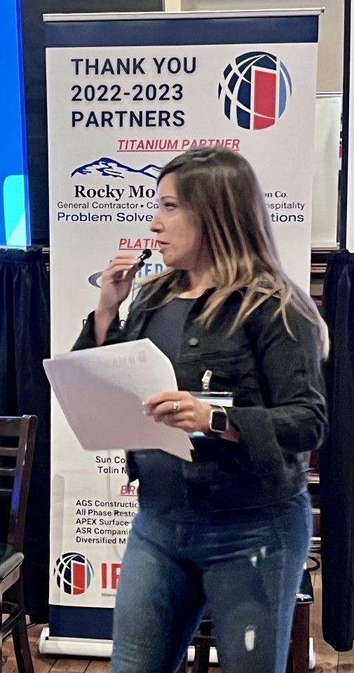 Woman speaking at event, holding papers and microphone, standing in front of a banner.