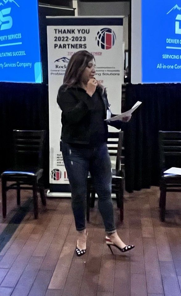 Woman speaking at an event, holding notes, wearing black and jeans, standing near a banner and chairs.