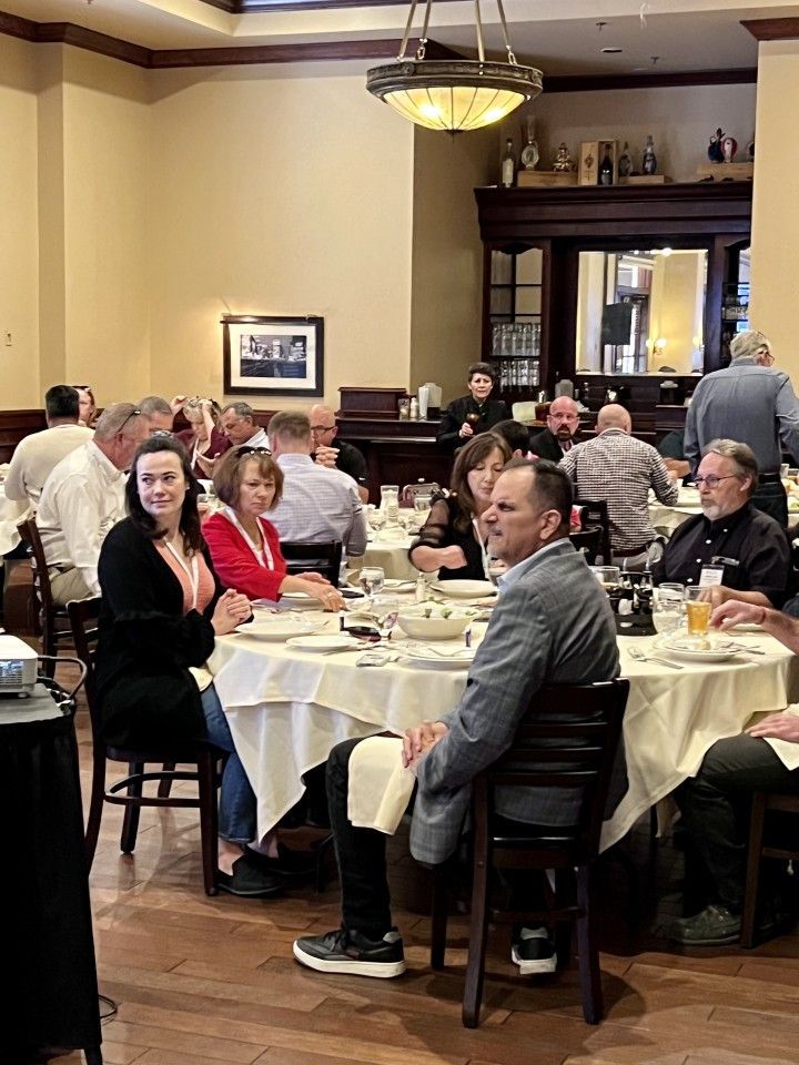 People seated at tables in a restaurant; some looking toward the camera, others talking. White tablecloths, wood paneling.