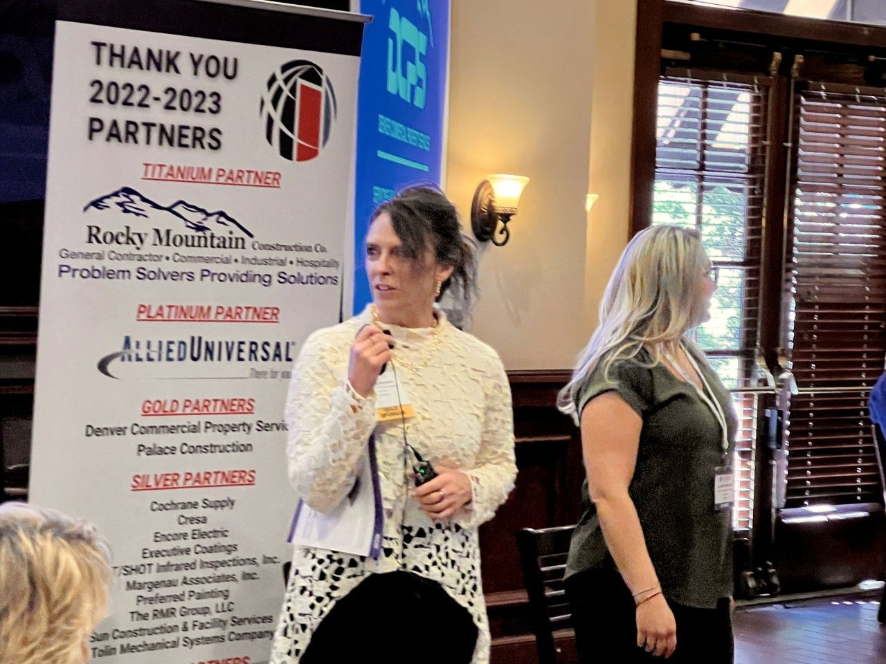 Two women at an event with a banner listing partners. One woman holds a bottle, speaks, and the other stands nearby.