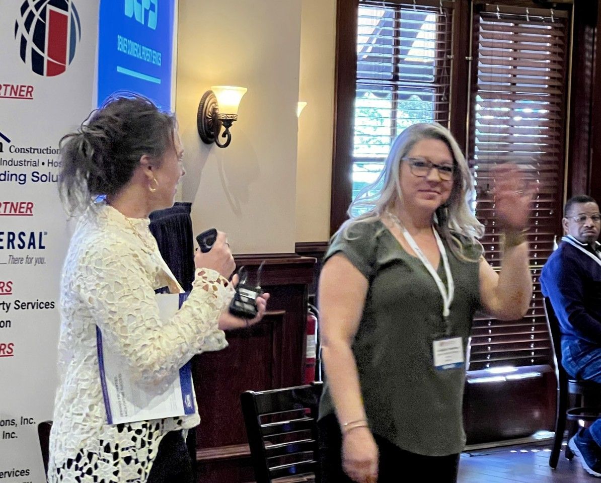 Woman with microphone gestures toward woman waving at event. Indoors, banner, chairs, window.