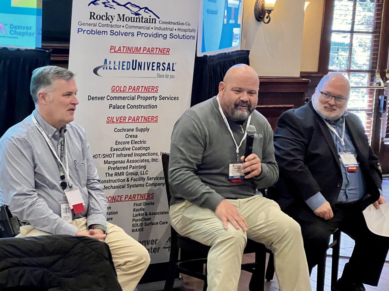 Three men seated at a panel, holding microphones. Banner behind them reads 