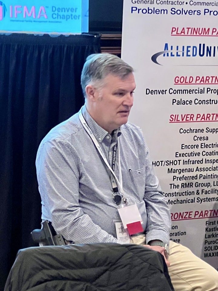 Man speaking at a conference. He wears a light-colored shirt and has a lanyard. Sponsors' names are on a banner behind him.