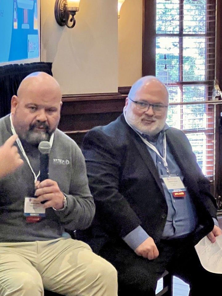 Two men seated at a panel, one speaking into a microphone. Both wear lanyards and are indoors.