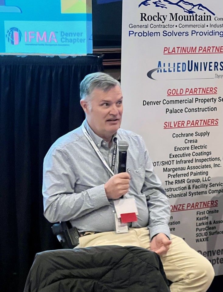 Man speaking into a microphone at an event, seated in front of a banner.
