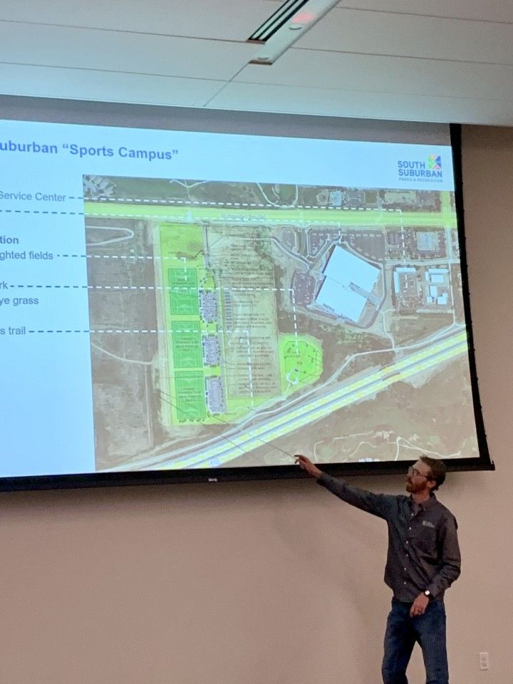 Man points to a presentation screen showing a sports complex layout, fields, and a building.