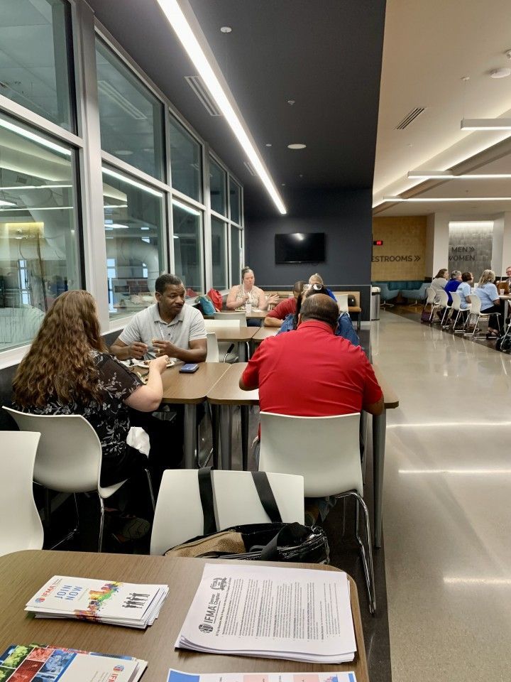 People seated at tables in a bright room with large windows, some looking at papers, others conversing.
