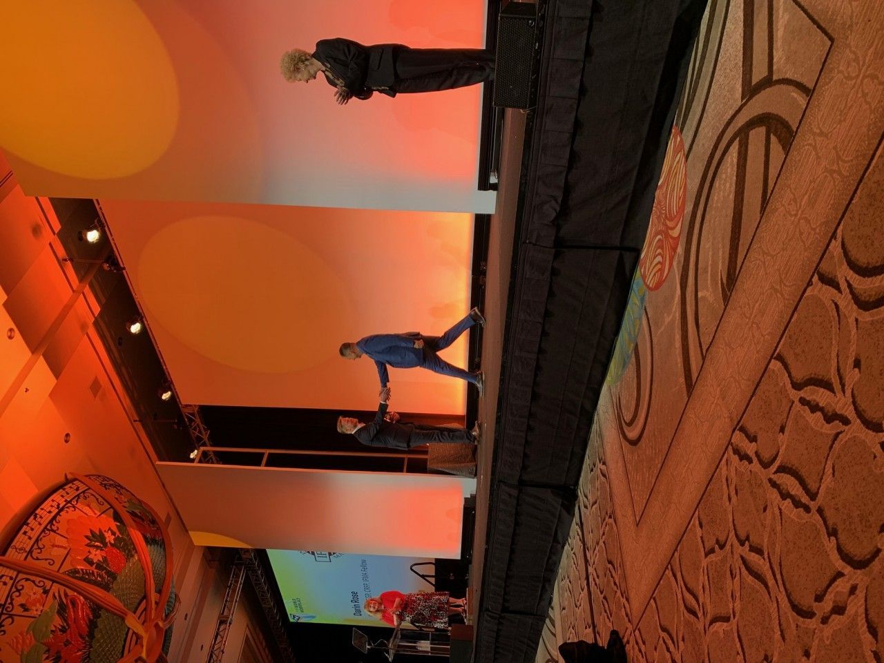 People on stage with an orange backdrop. One person is balancing, while the others are standing.