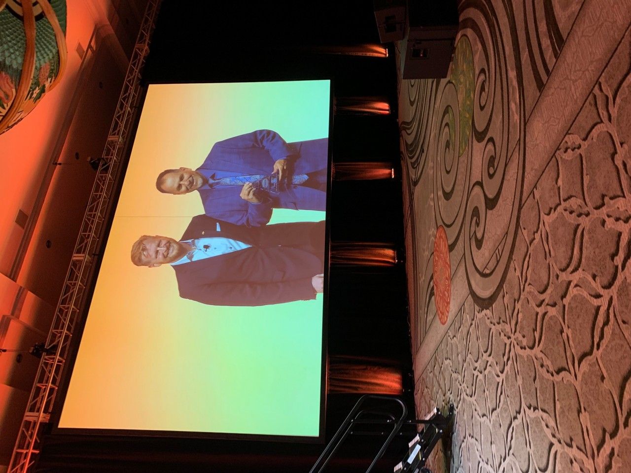 Two men on a screen during an event; one in a blue blazer, the other in a black blazer. The setting appears to be a stage.