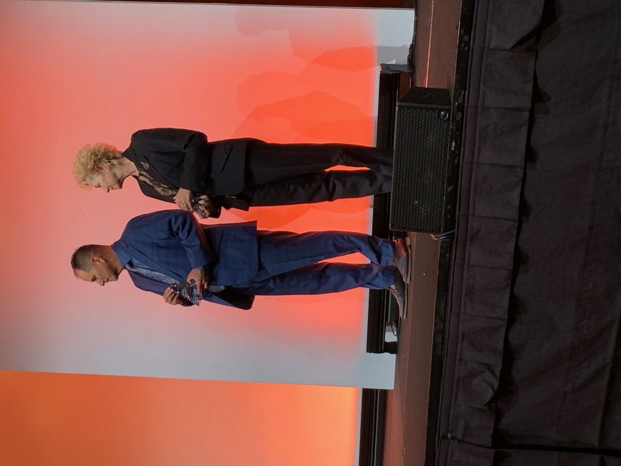 Two people on stage: man in blue suit, woman in black suit, facing a white and orange backdrop.
