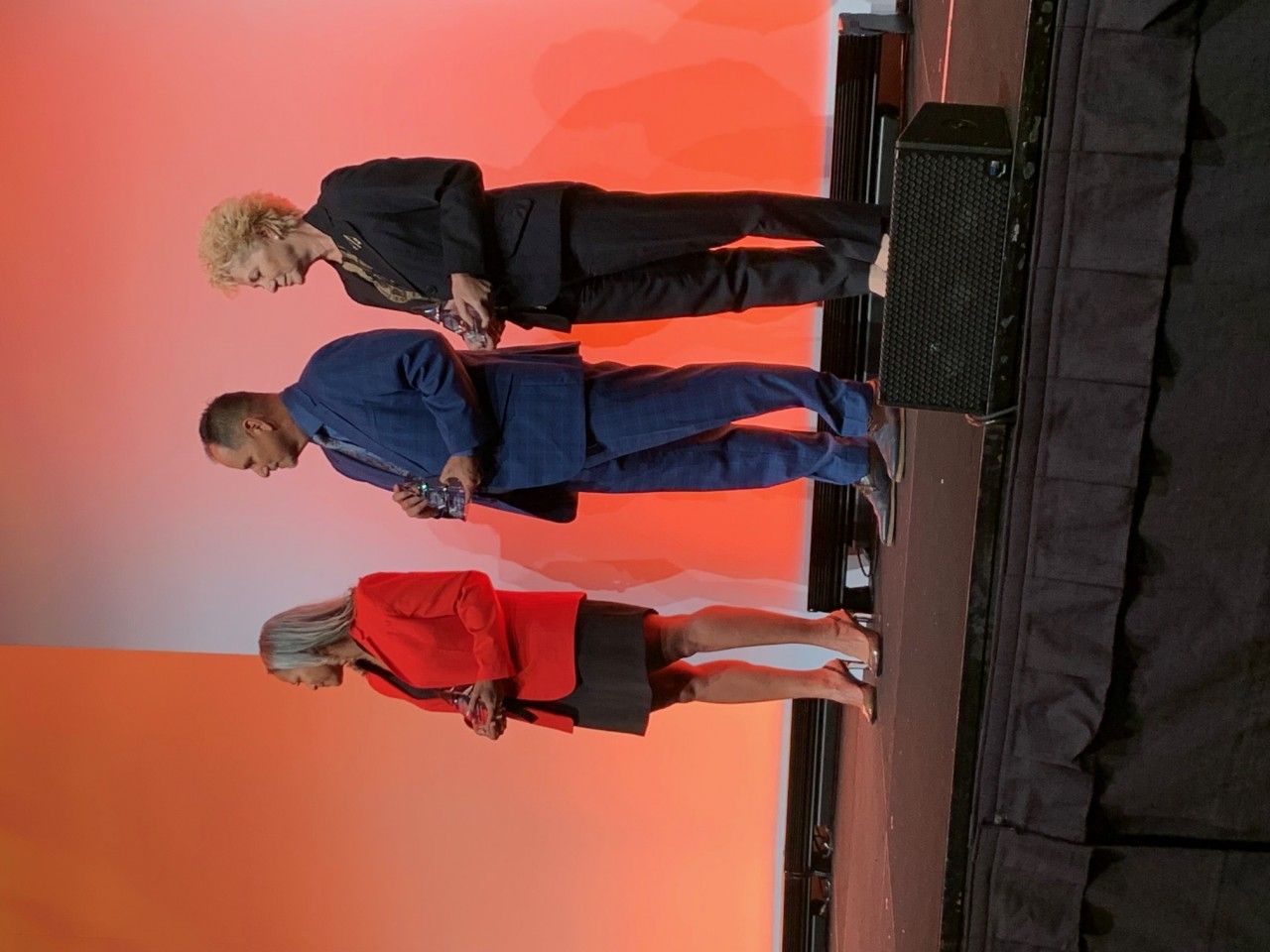 Three people standing on a stage with an orange backdrop. The people appear to be presenting.