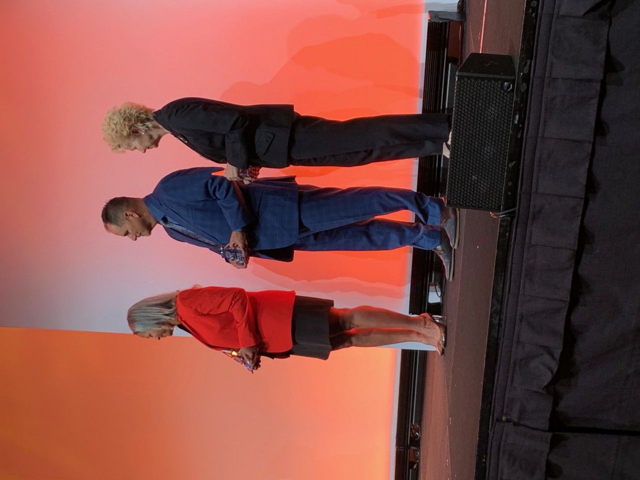 Three people in suits bowing on stage. Orange backdrop.