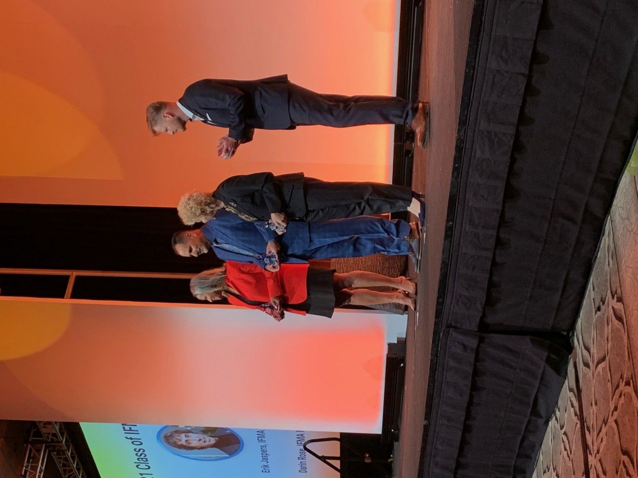 People on stage, one in a suit bowing, others standing. Orange background.