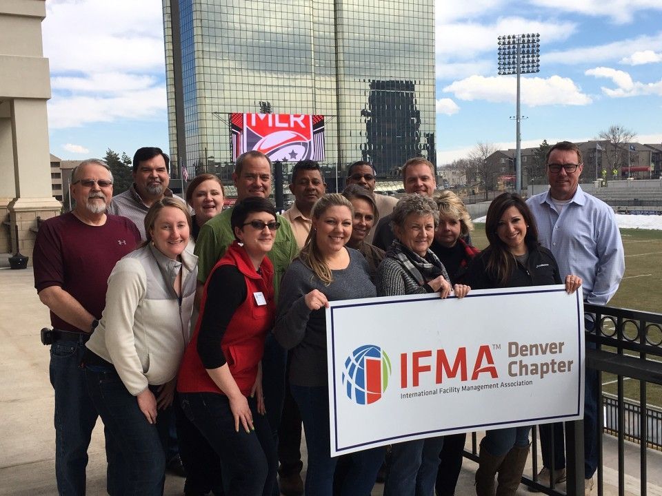 Group of people holding an IFMA Denver Chapter sign in front of a stadium and a mirrored building.