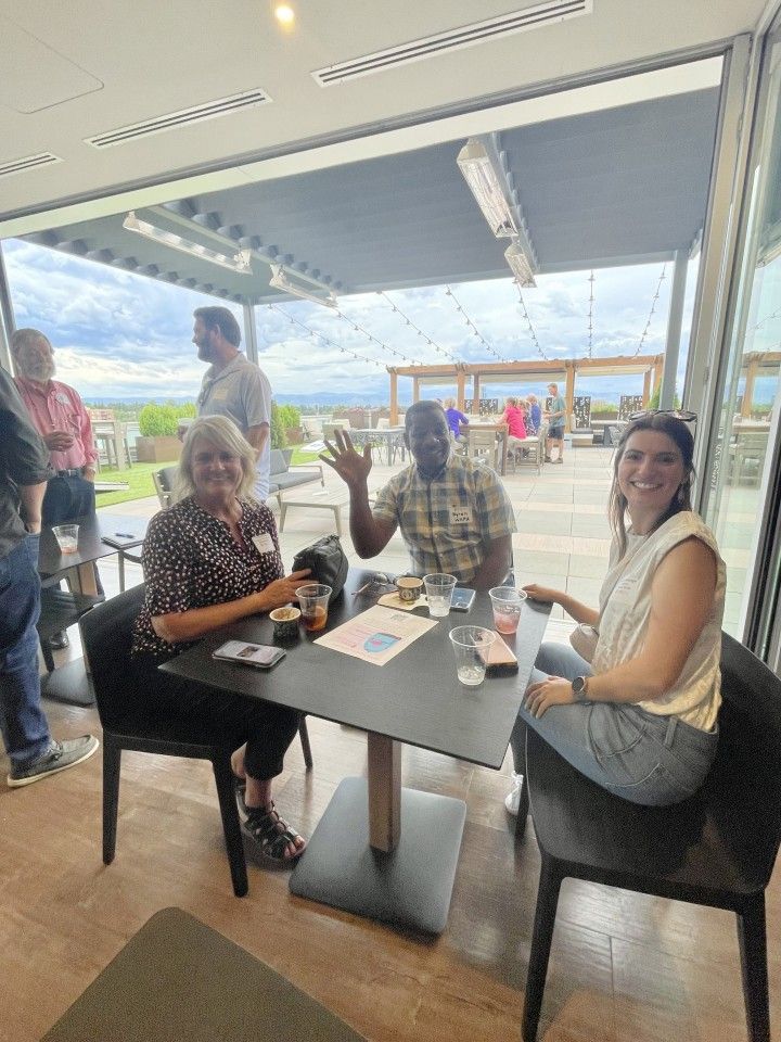 Three people at a table, smiling and waving, inside a venue with an outdoor view.