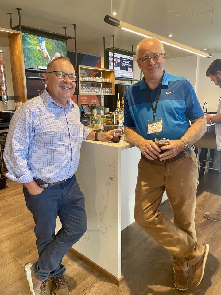 Two men at a bar, leaning casually. One in blue shirt, other in light blue plaid shirt. Bar setting, light wood floors.