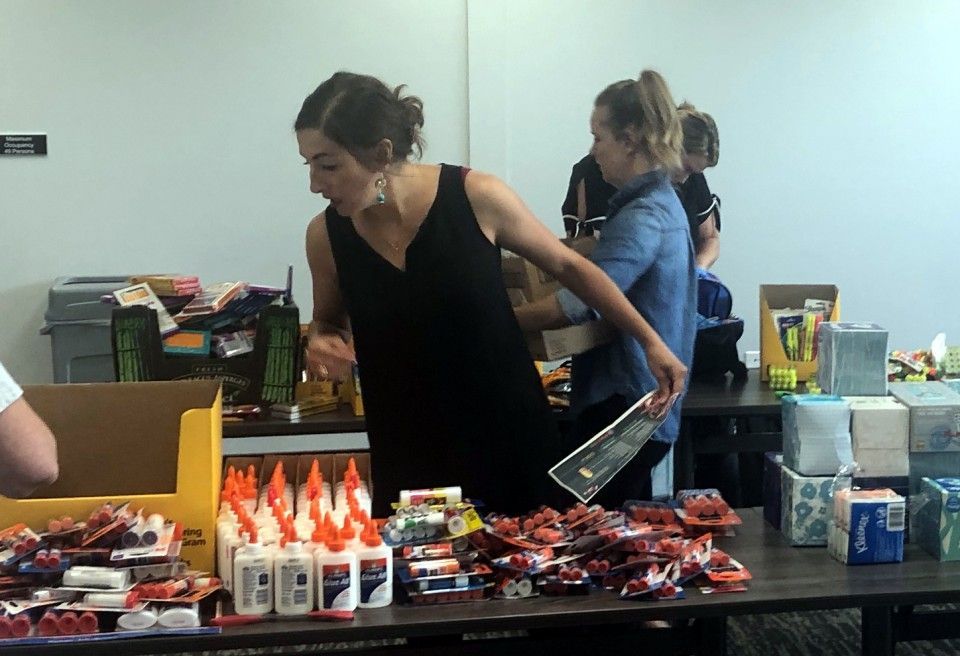 People organizing school supplies on a table; woman in black dress, glue bottles, and boxes of items.