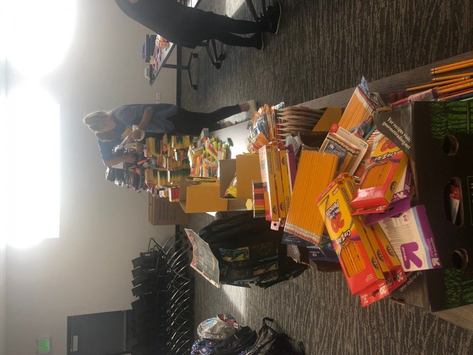 People sorting school supplies on a table in a room. Pencils, erasers, and notebooks are visible.