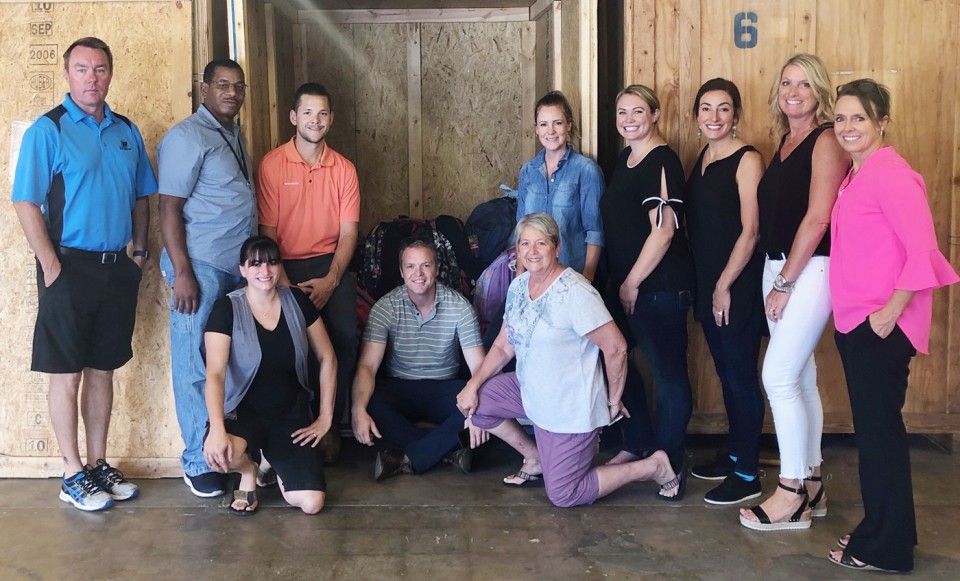 Group of people pose in front of a storage unit, some kneeling, smiling. Wooden walls.