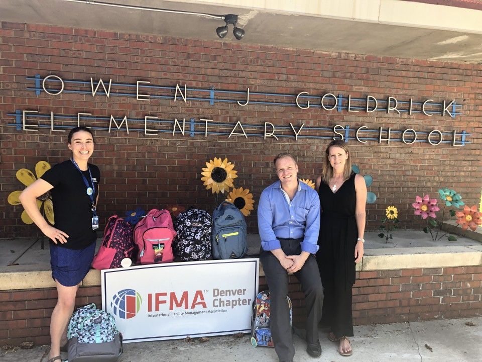 People stand with backpacks in front of Owens J. Goldrick Elementary School, Denver. IFMA Denver Chapter logo visible.