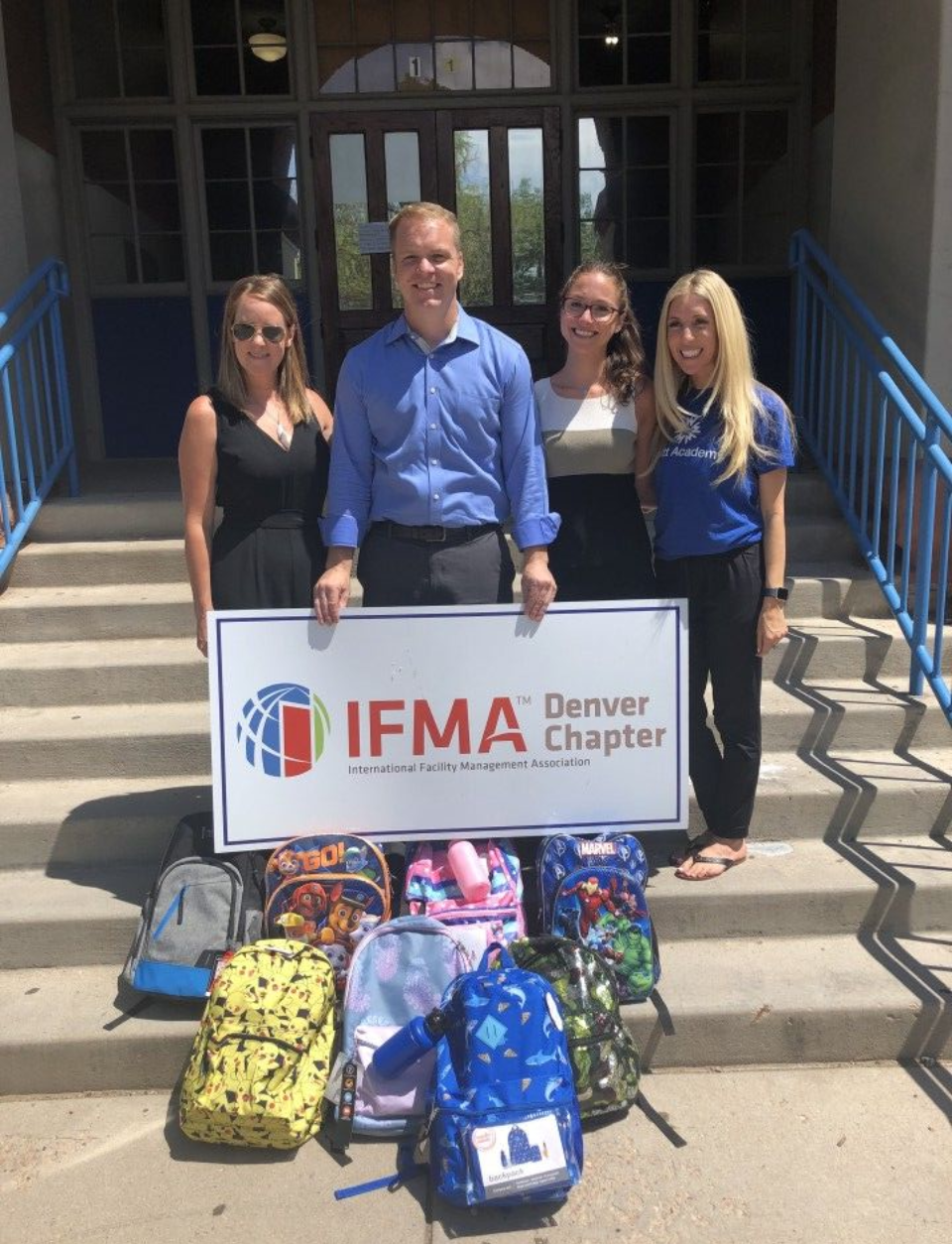 Four people stand outside a building, holding an IFMA Denver Chapter sign with backpacks.