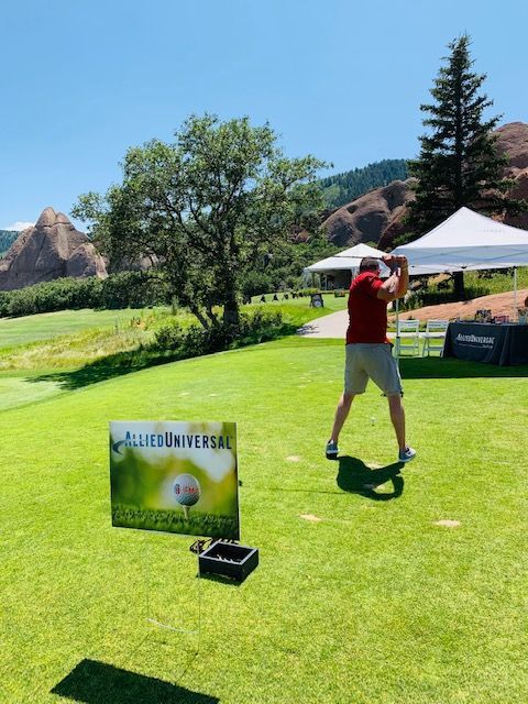 Golfer swinging on a green golf course, sign nearby. Sunny day, red shirt, beige shorts.