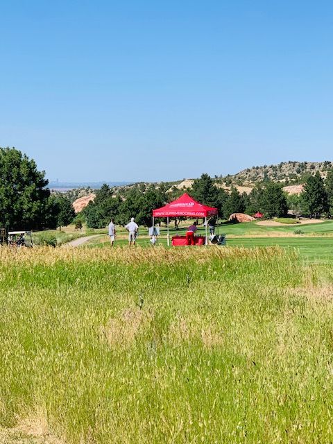 Green golf course with people near a red tent, under a blue sky, on a sunny day.