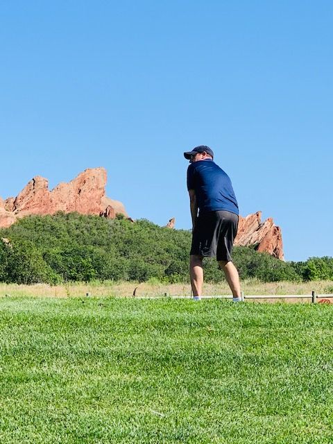 Golfer taking a swing on a green course, with red rock formations in the background. Bright sunny day.