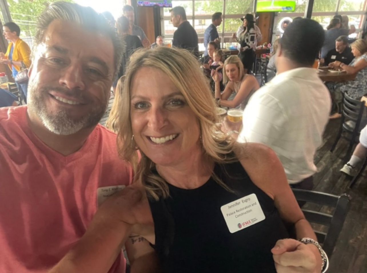 Man and woman smiling, fist bumping at an outdoor gathering, other people in the background.