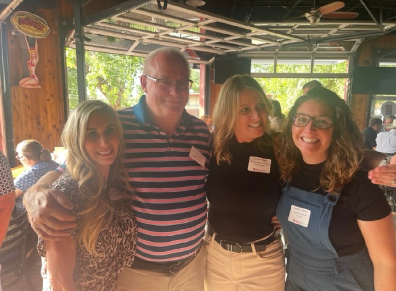 Four people smiling, posing together inside a wood-paneled building with overhead fans and natural light.
