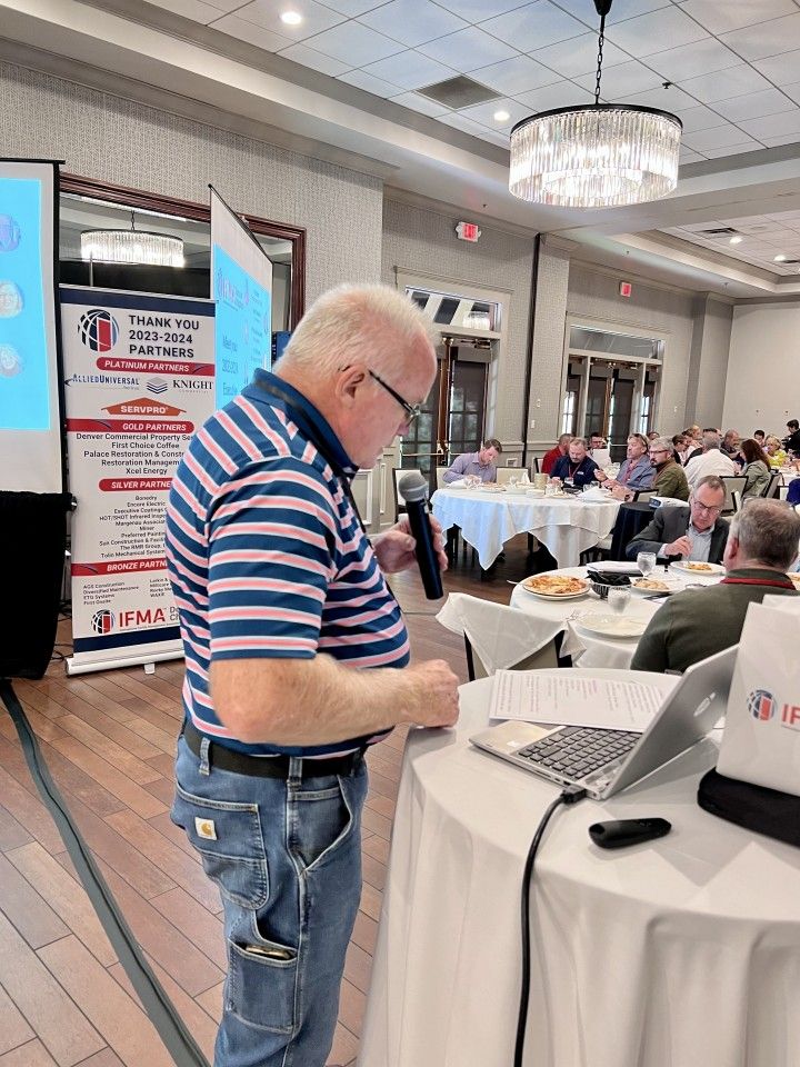 Man in striped shirt at a podium, speaking at a conference. Others seated at tables.