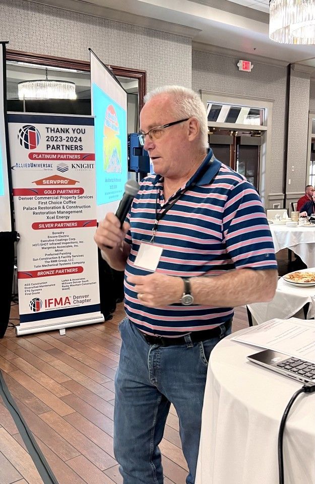 Man speaking into a microphone, standing near a banner at an event. He wears a striped shirt and jeans.