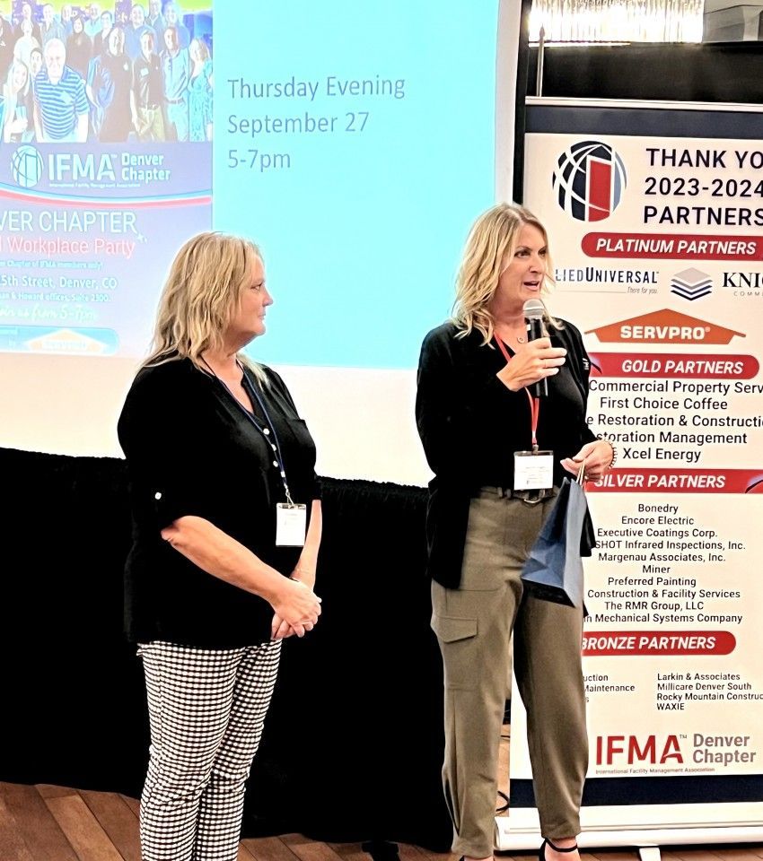 Two women speaking at a conference, IFMA Denver Chapter. One holds a microphone. Sponsors' logos in the background.