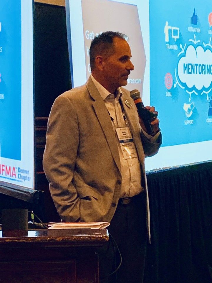Man in blazer speaking into microphone at conference, Mentoring graphic in background.