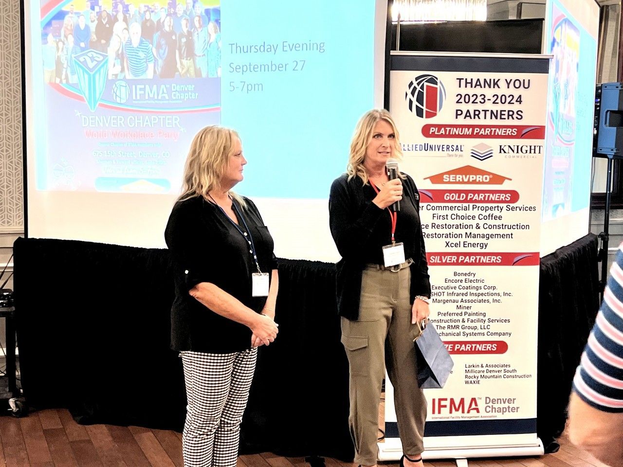 Two women at a podium speaking at a conference. A banner and screen with text are behind them.