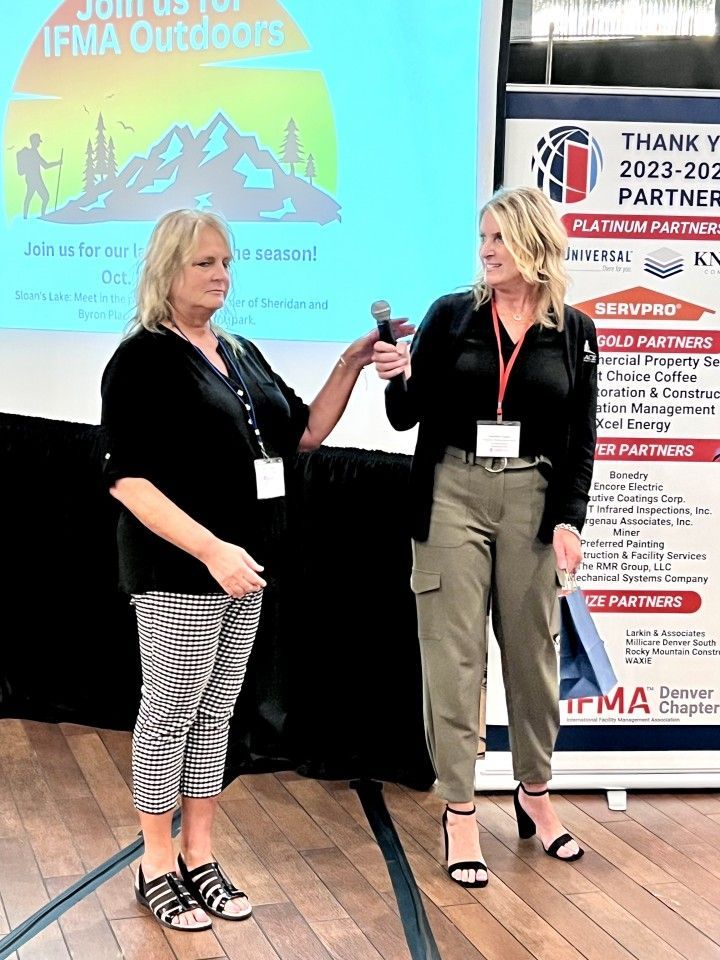 Two women on stage, one holding a microphone, appearing to speak at an IFMA Outdoors event.