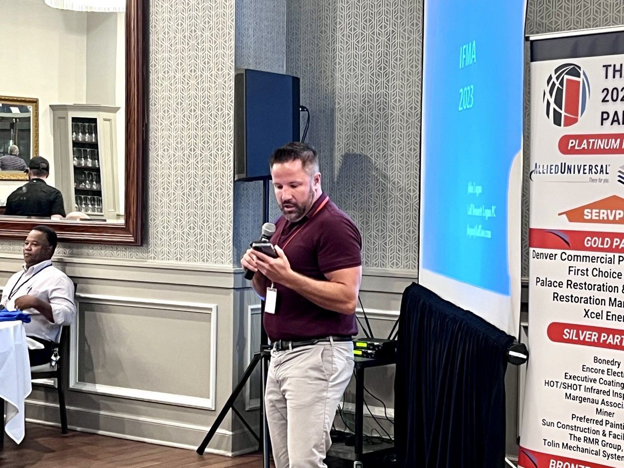 Man speaking at a podium, holding phone. Banner behind him. Other people seated.