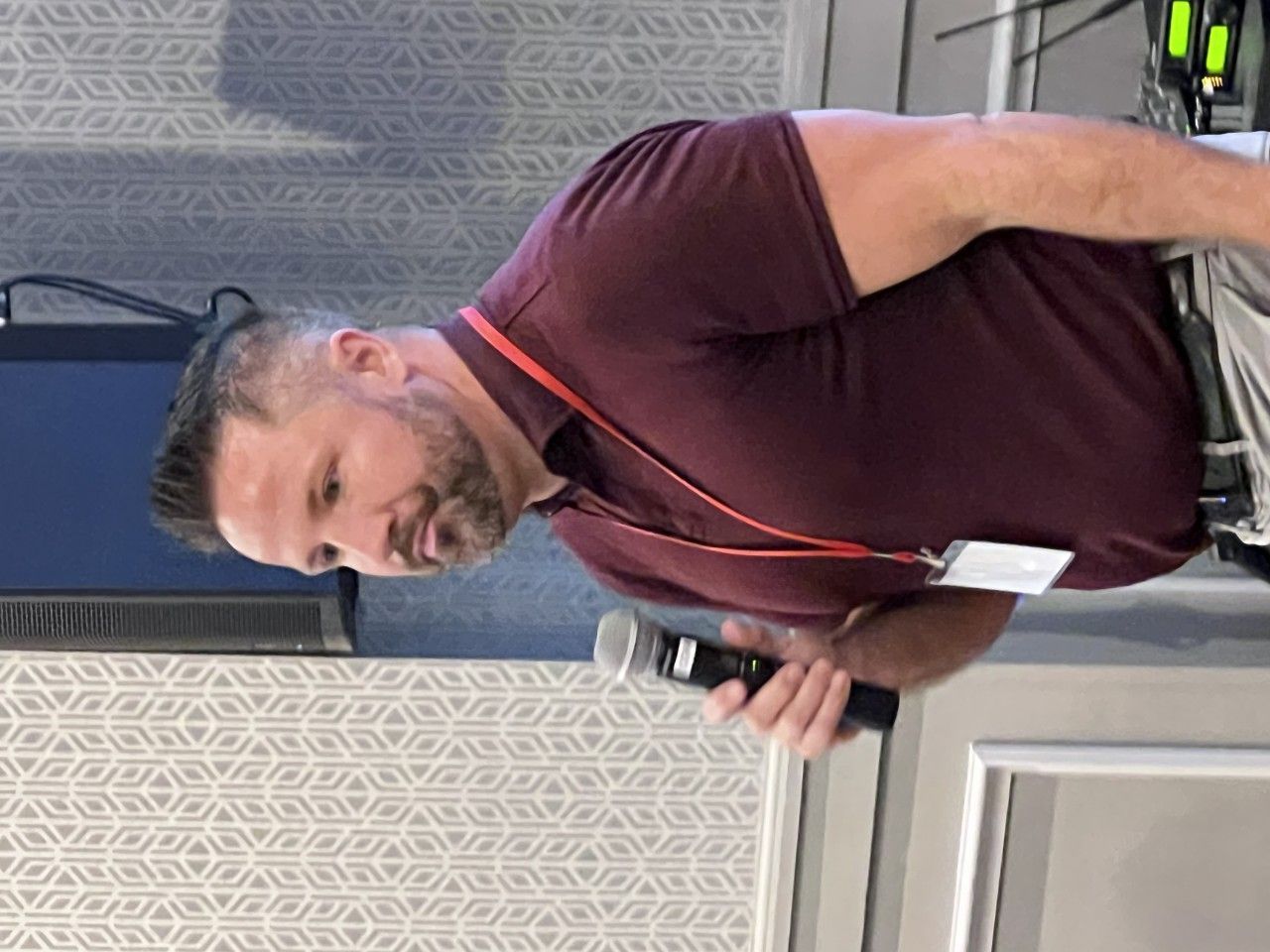 Man in maroon shirt speaking into a microphone at a conference, holding a name tag.