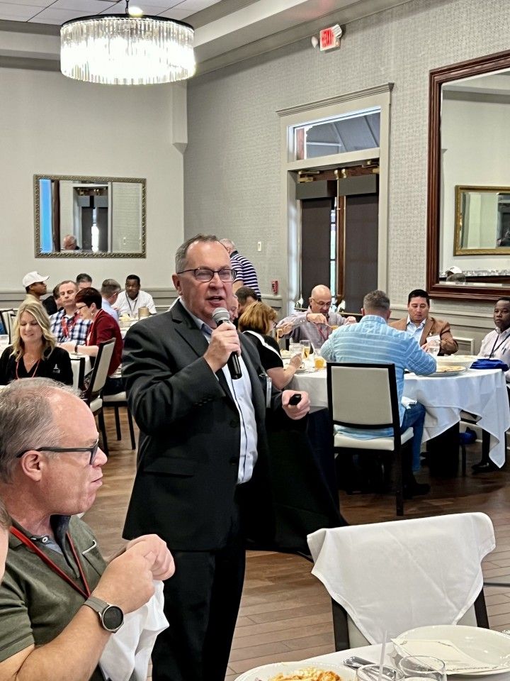 Man speaking into a microphone at a formal event, tables with seated guests in background.