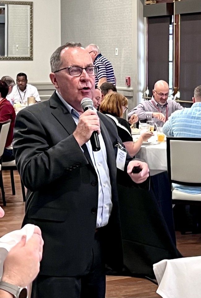 Man speaking into microphone at a conference; other attendees at tables in the background.