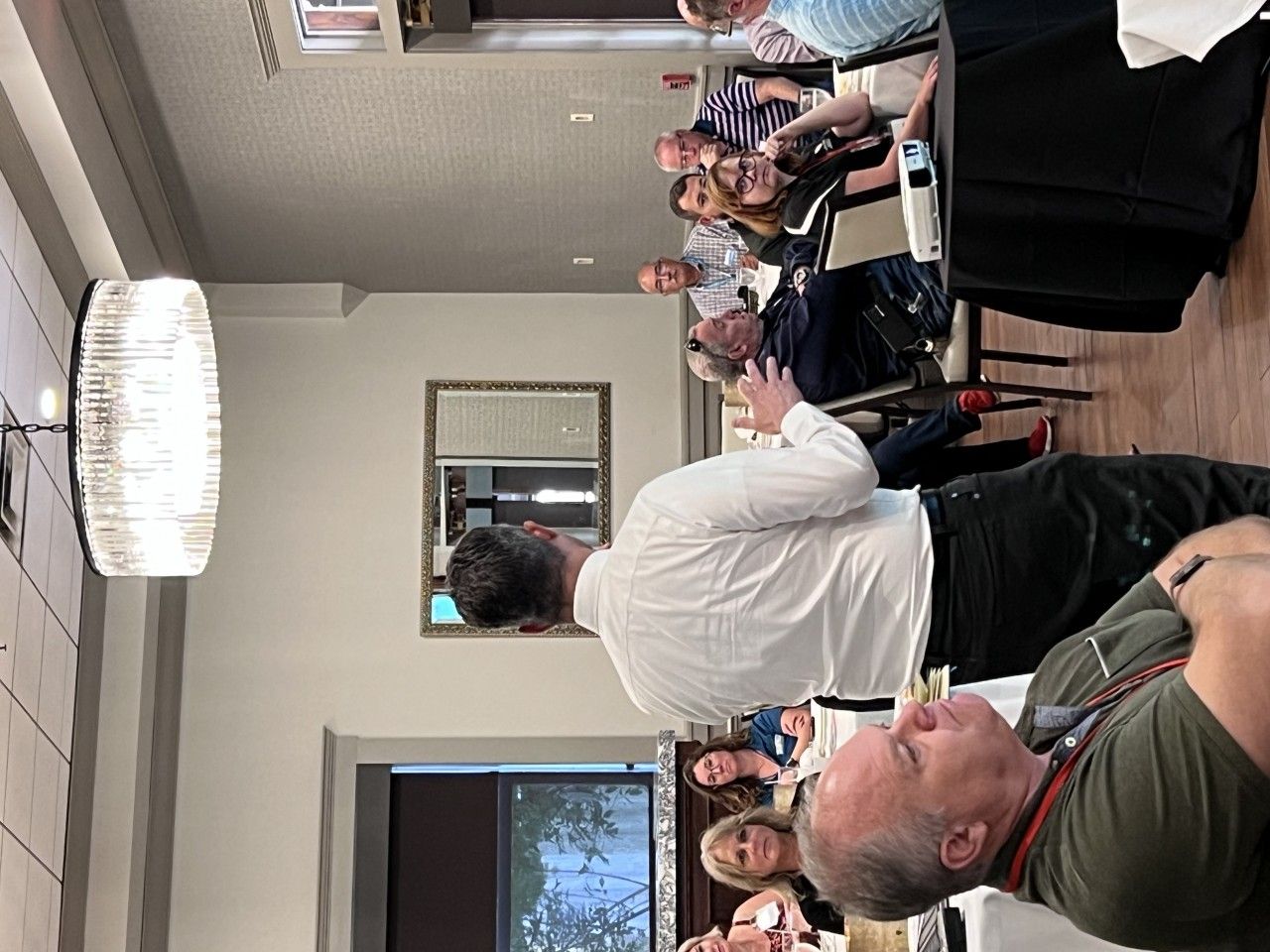 People at a meeting, man speaking, overhead light, others seated at tables.