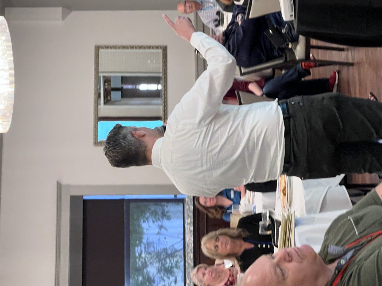 Man speaking, gesturing with raised arm, at a table with people in a well-lit indoor setting.