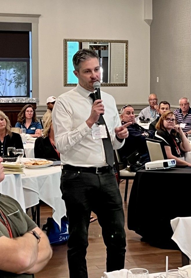 Man speaking into a microphone at an event, gesturing with his hand. Attendees are seated at tables in a room.