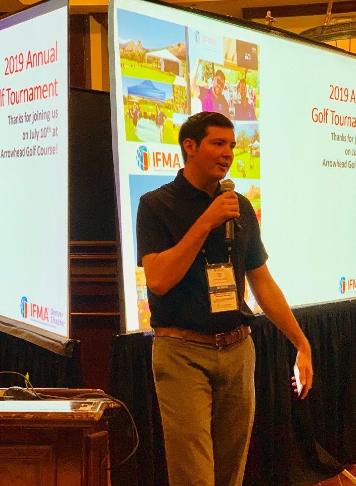 Man speaking at a conference, holding a microphone, with a screen in the background advertising a golf tournament.