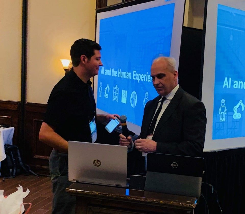 Two men at a conference; one shows a phone, the other holds a pen. A screen displays 