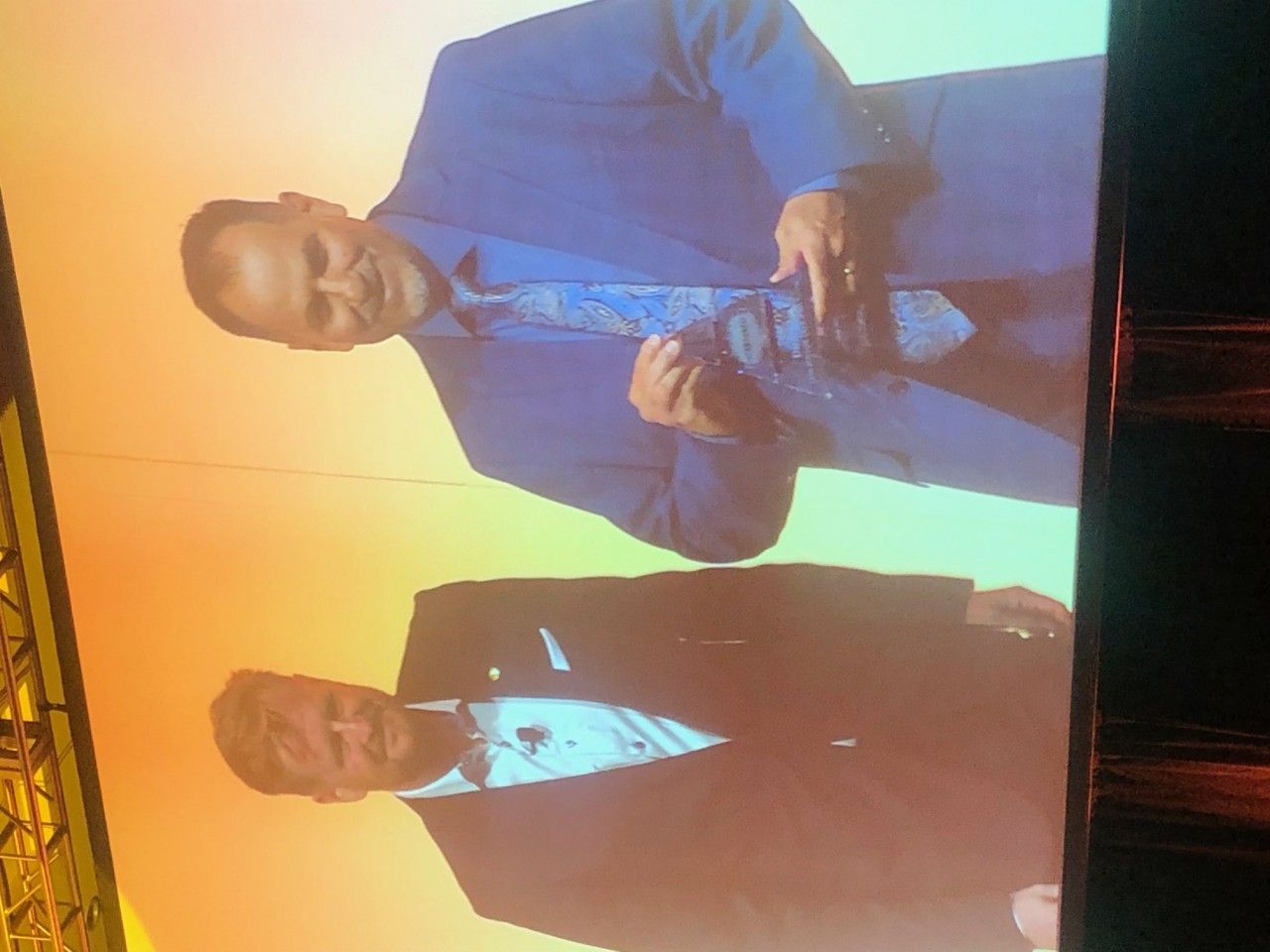 Two men on stage, one in blue suit adjusting tie, the other in black suit. Yellow-orange background.